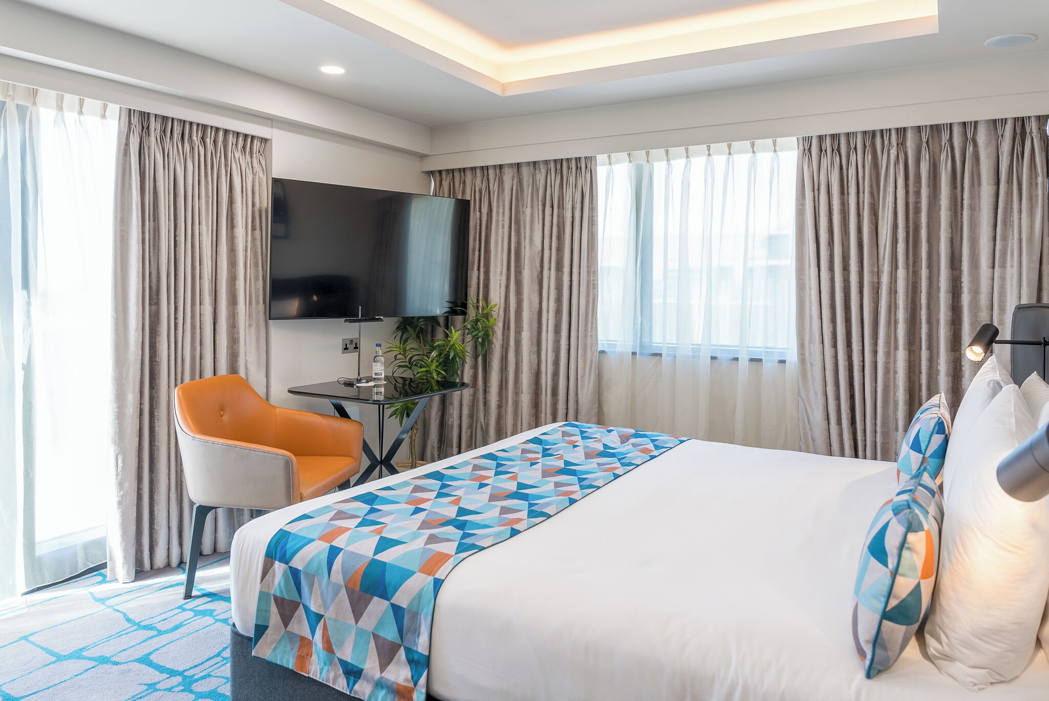A modern hotel room with a large bed, a desk with a flat-screen TV, an orange chair, and a potted plant.