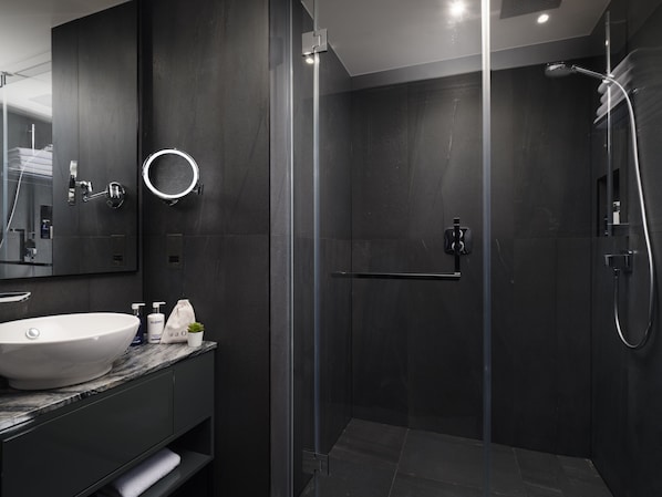 Um banheiro moderno com uma pia grande e branca, azulejos escuros, chuveiro com box de vidro e um espelho com iluminação circular.