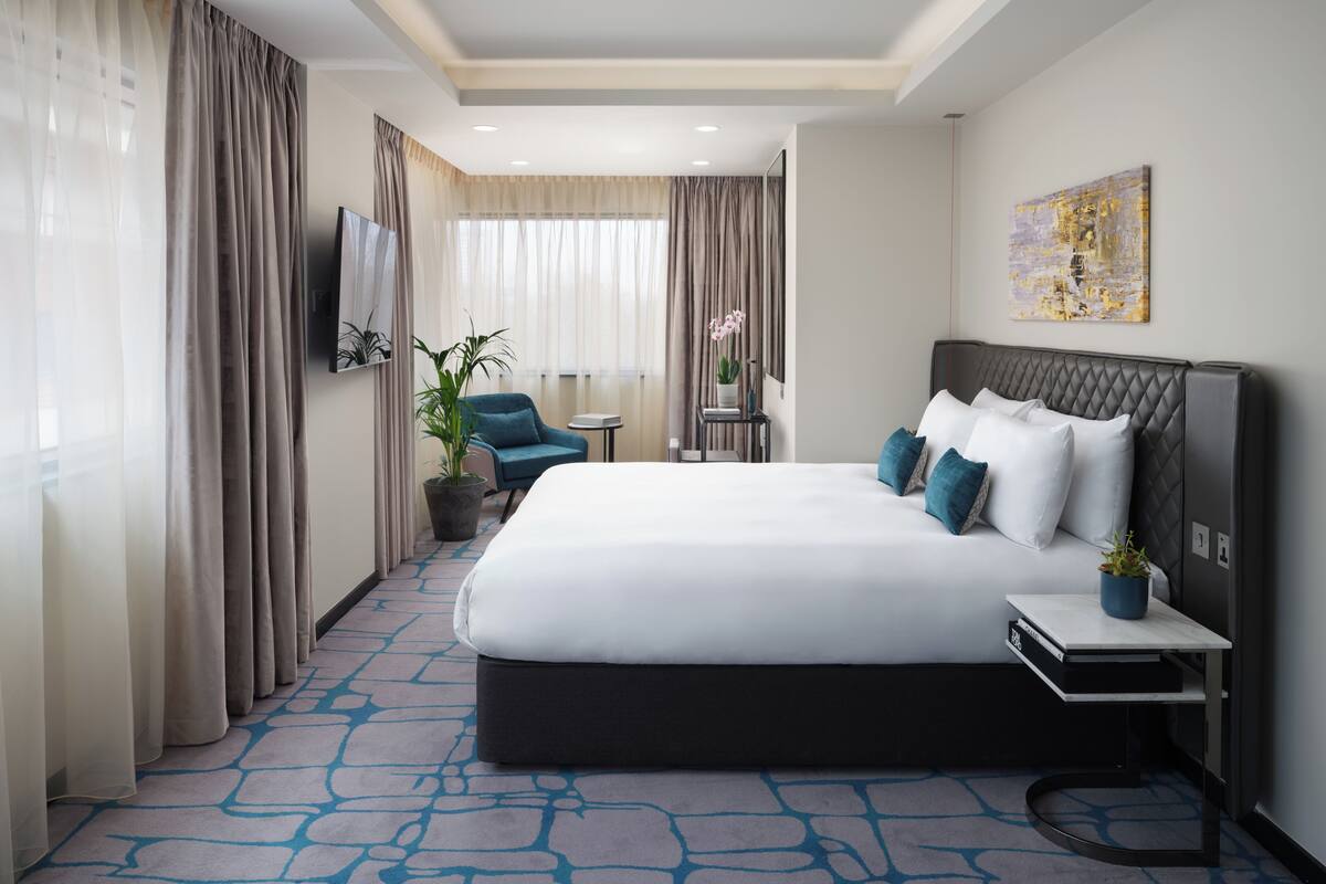 Une chambre d’hôtel avec un grand lit, un petit bureau, une chaise, une plante en pot et un tableau au mur.