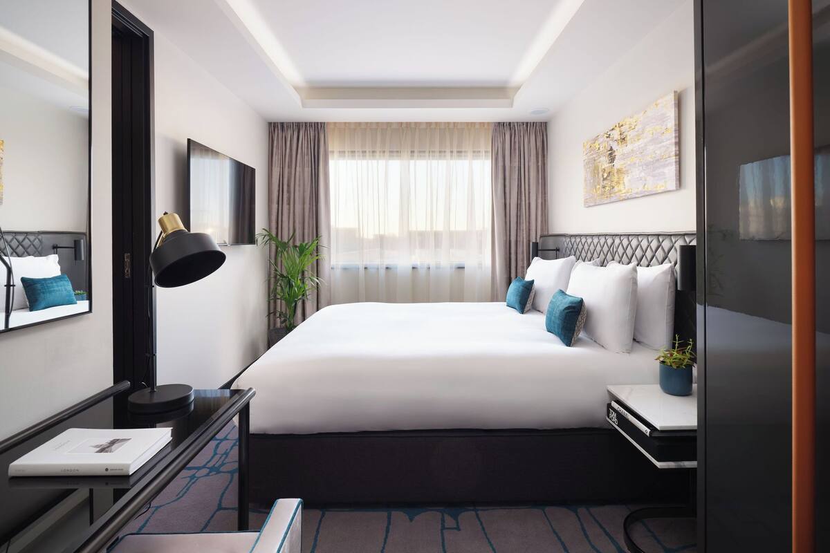 Une chambre d’hôtel moderne avec un grand lit, un bureau avec une lampe, une plante en pot et une fenêtre avec des rideaux.