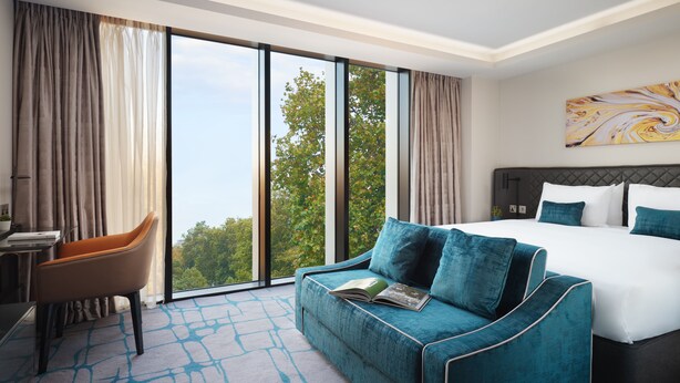A hotel room with a large bed, a sofa, a desk, and a view of trees through large windows.