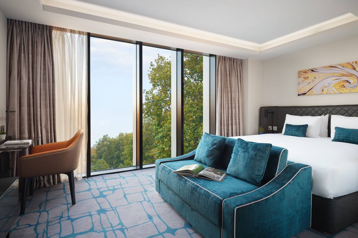 Une chambre d’hôtel avec un grand lit, un canapé, un bureau et une vue sur les arbres, visible à travers de grandes fenêtres.