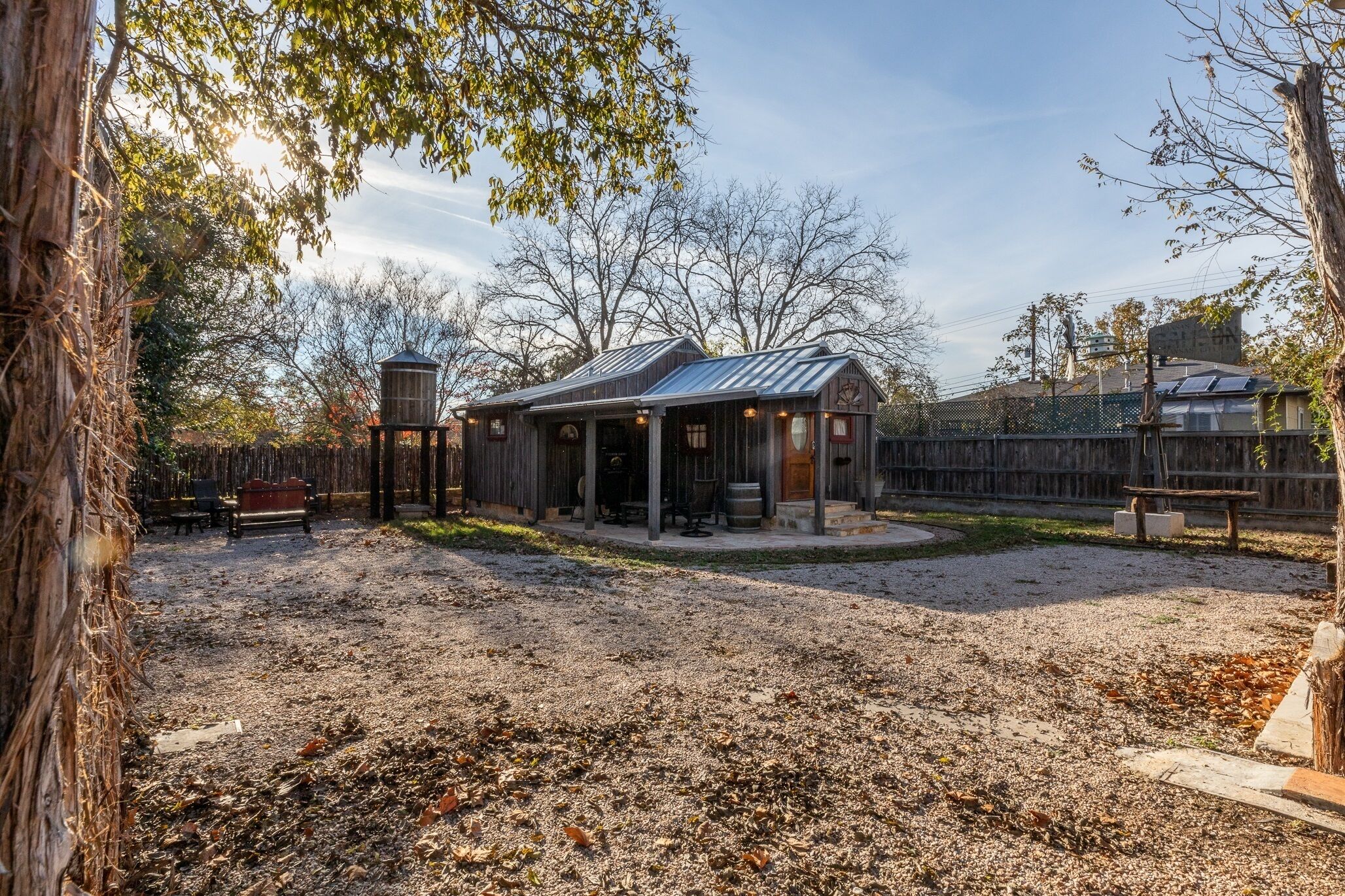 A rustic cabin with a metal roof, surrounded by a fenced yard with gravel paths and leaf-covered ground.