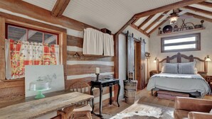A rustic room with a wooden ceiling, a bed with a wooden headboard, a table with chairs, and a window with curtains. - Cozy Cabin for Two 1 Block to Main (Fredericksburg)