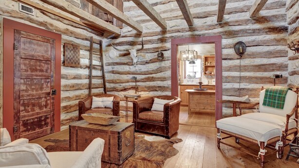 Interior rústico de una cabaña de troncos con muebles de cuero, una cama de madera y una pared de piedra decorada con un cráneo de vaca.