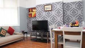 Uma sala de estar com um sofá, uma televisão sobre um móvel, uma mesa de jantar com cadeiras e uma fruteira.