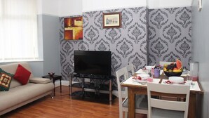 Uma sala de estar com sofá, televisão em móvel, mesa de jantar com cadeiras e uma fruteira.