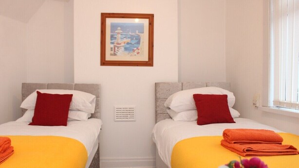 Quarto de hotel com duas camas, cada uma com uma almofada vermelha e um cobertor amarelo. Há um quadro na parede acima das camas.
