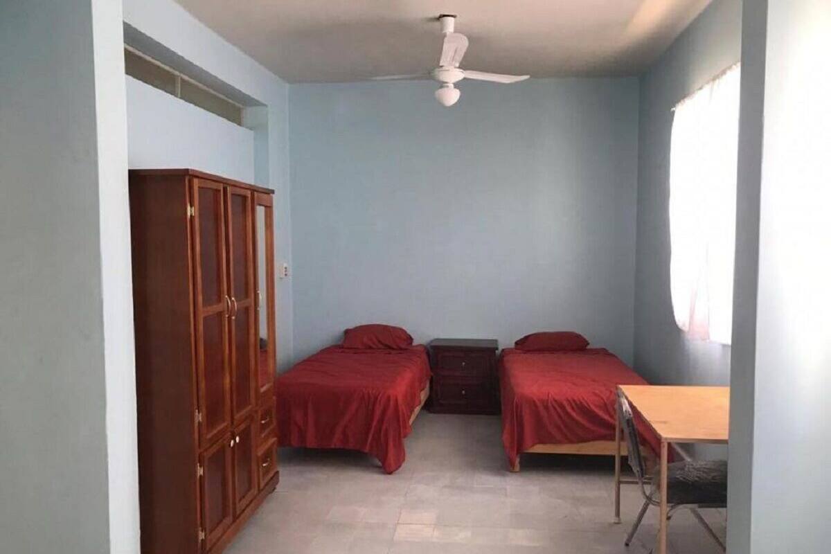 A small, simple room with two single beds, a wooden wardrobe, a desk, and a ceiling fan.