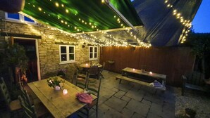 Une terrasse extérieure avec des murs en pierre, des guirlandes lumineuses et une table en bois entourée de chaises.