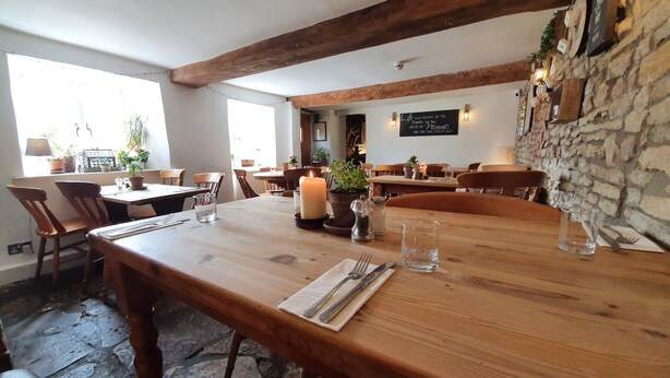Un coin repas rustique avec des tables et des chaises en bois, un mur en pierre et un centre de table composé de bougies.
