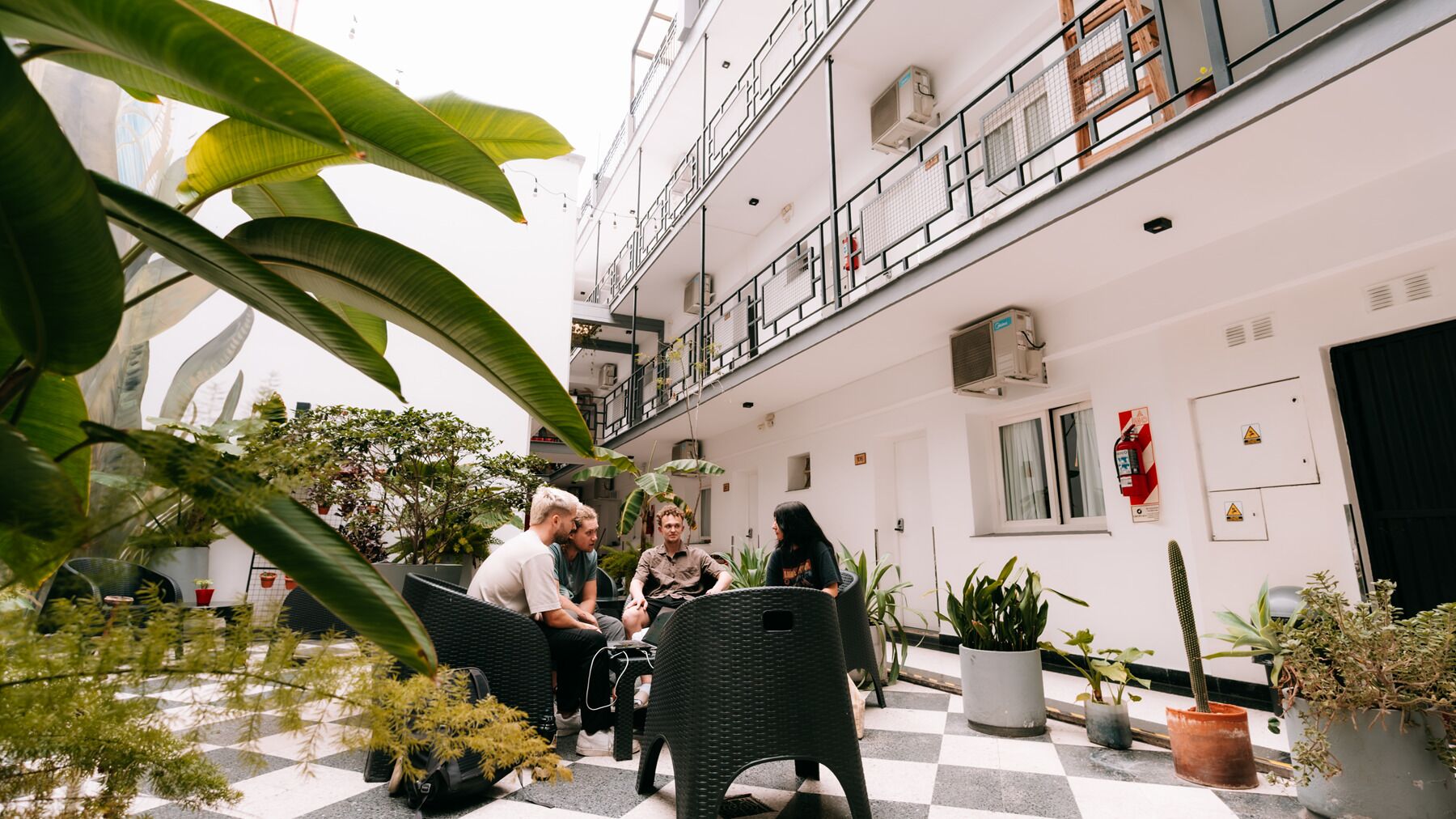 A multi-level indoor space with a checkered floor, potted plants, and a group of people seated on wicker furniture.