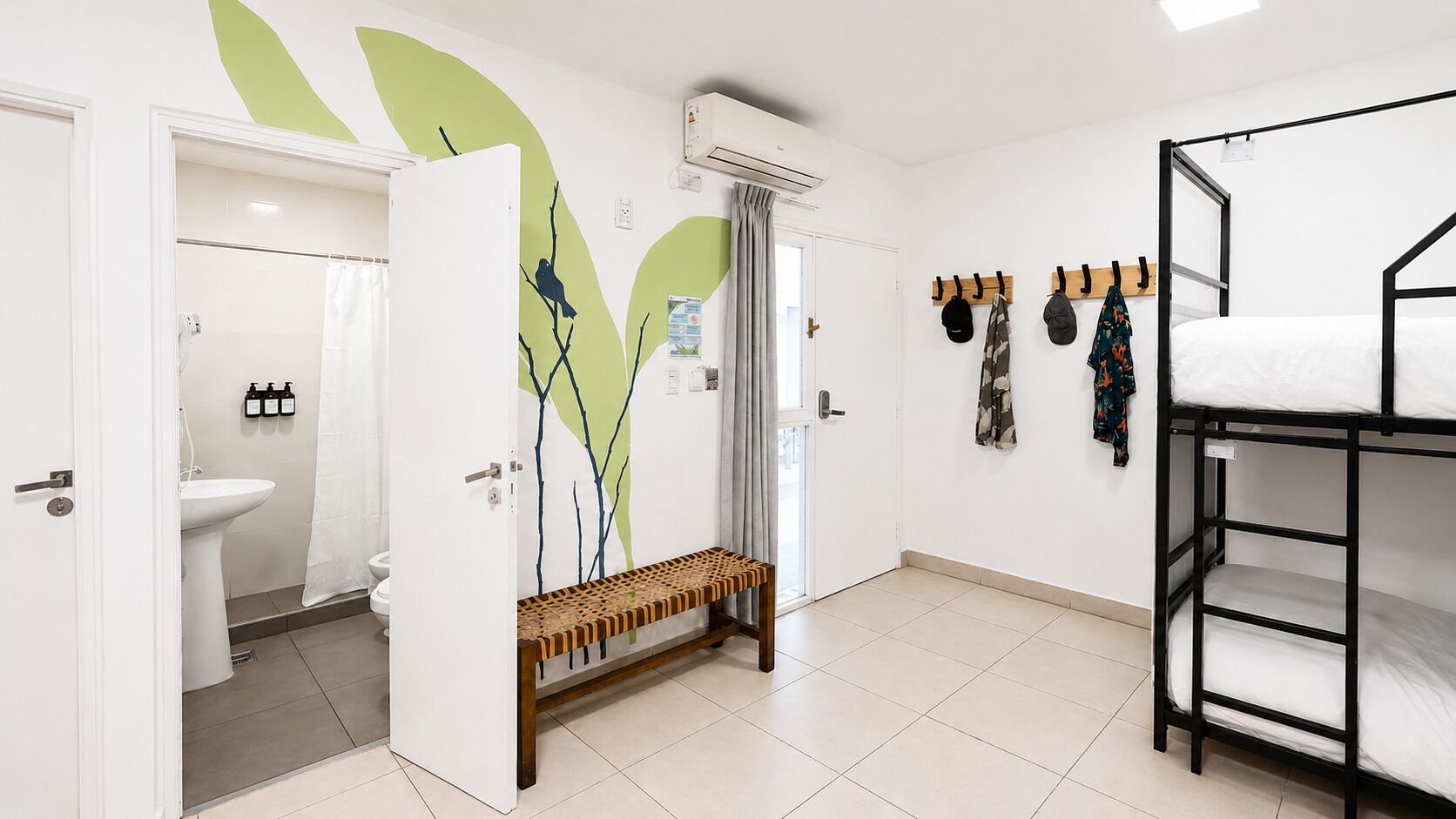 Una habitación con literas, un banco, un perchero en la pared y un baño con inodoro y lavamanos.