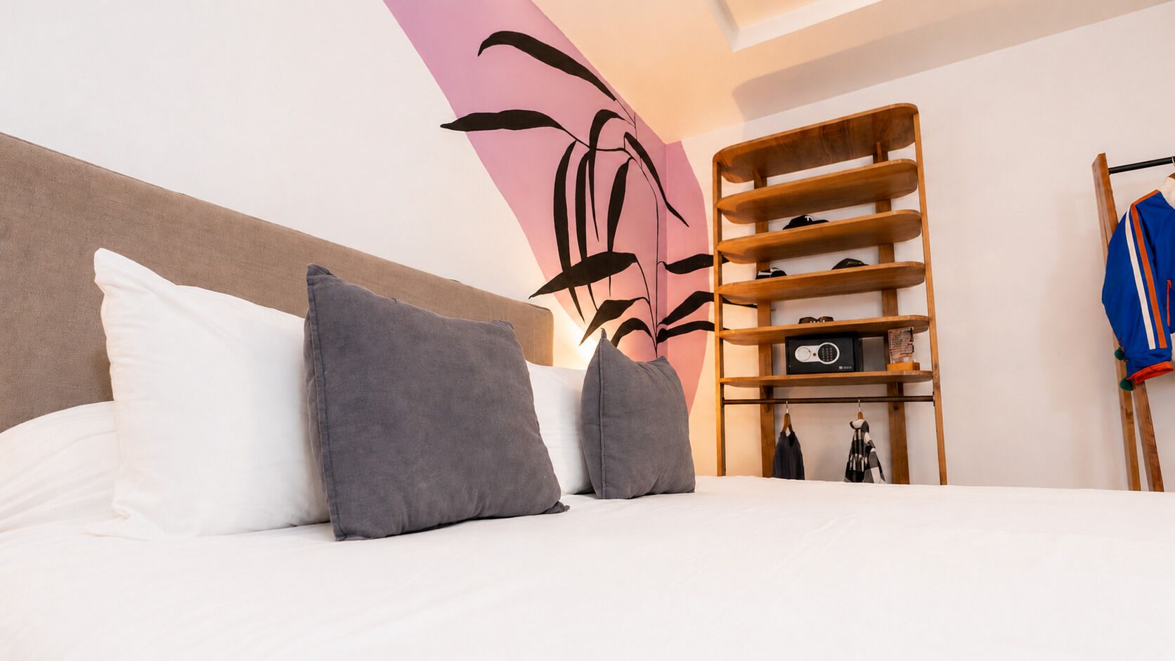 Habitación de hotel con cama, almohadas, una obra de arte en la pared, un estante de madera con objetos y un perchero.