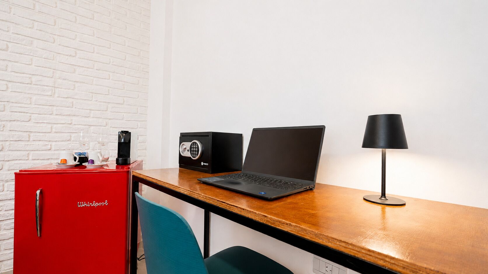 A red Whirlpool mini-fridge, a black laptop, a black speaker, and a black desk lamp on a wooden desk against a white brick wall.
