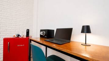 A red Whirlpool mini-fridge, a black laptop, a black speaker, and a black desk lamp on a wooden desk against a white brick wall.