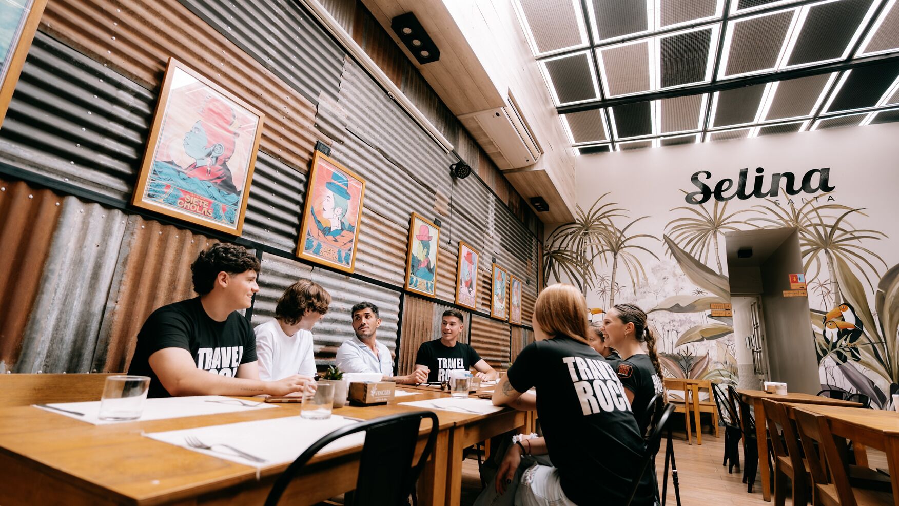 A group of people seated at a wooden table in a restaurant with a corrugated metal wall and framed artwork.