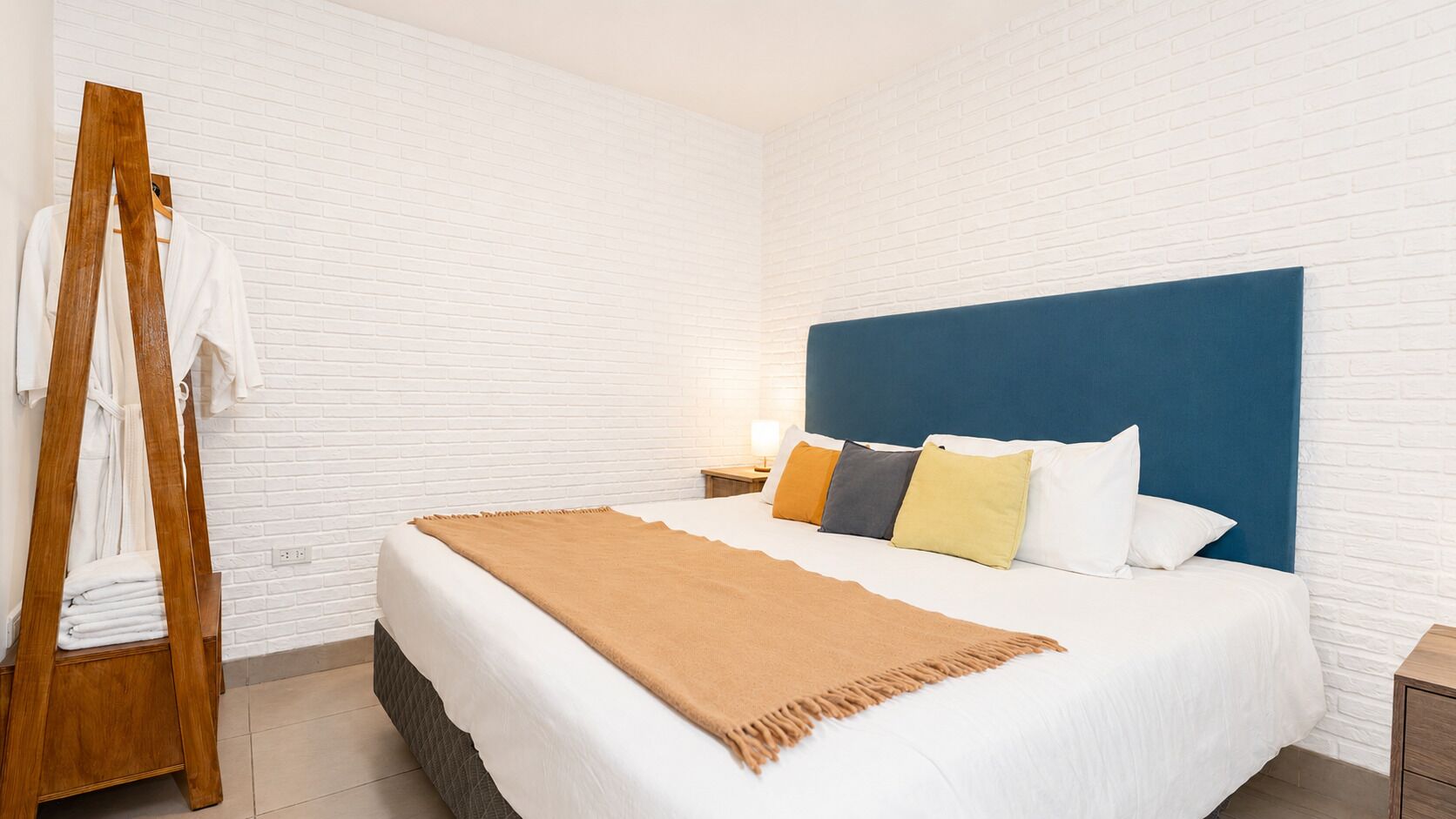 Habitación de hotel con pared de ladrillo blanco, cama con cabecera azul y un soporte de madera con toallas.