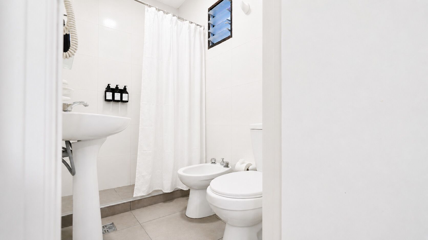 Une salle de bain avec un lavabo blanc, deux toilettes, un rideau de douche et un miroir.