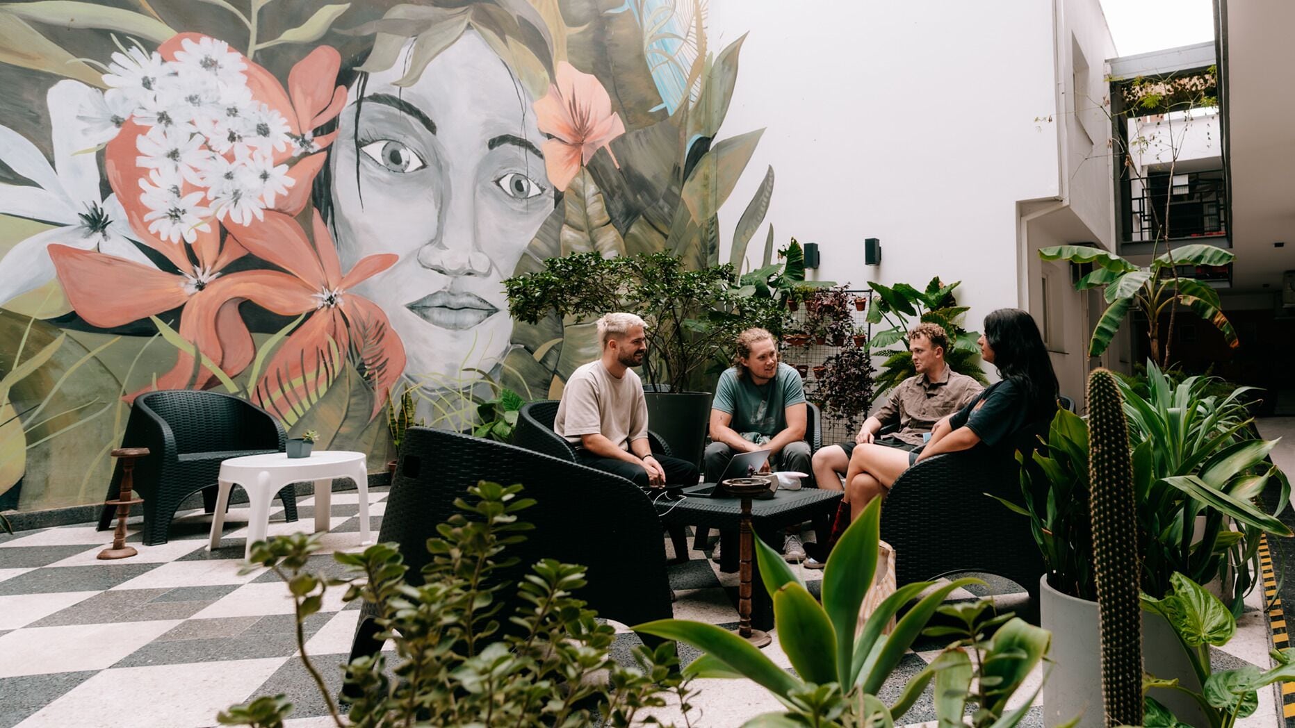 A group of people seated in a modern outdoor lounge area with a large mural, surrounded by various plants and furniture.