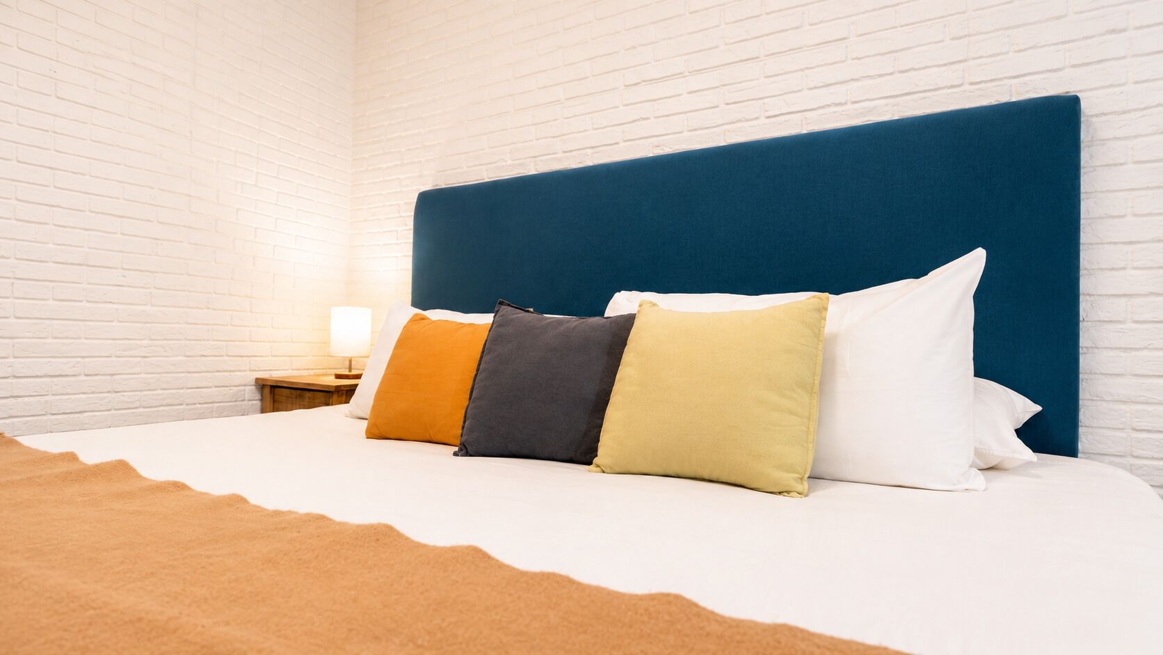 Habitación de hotel con pared de ladrillo blanco, cabecera azul y cama con almohadas blancas y de colores.