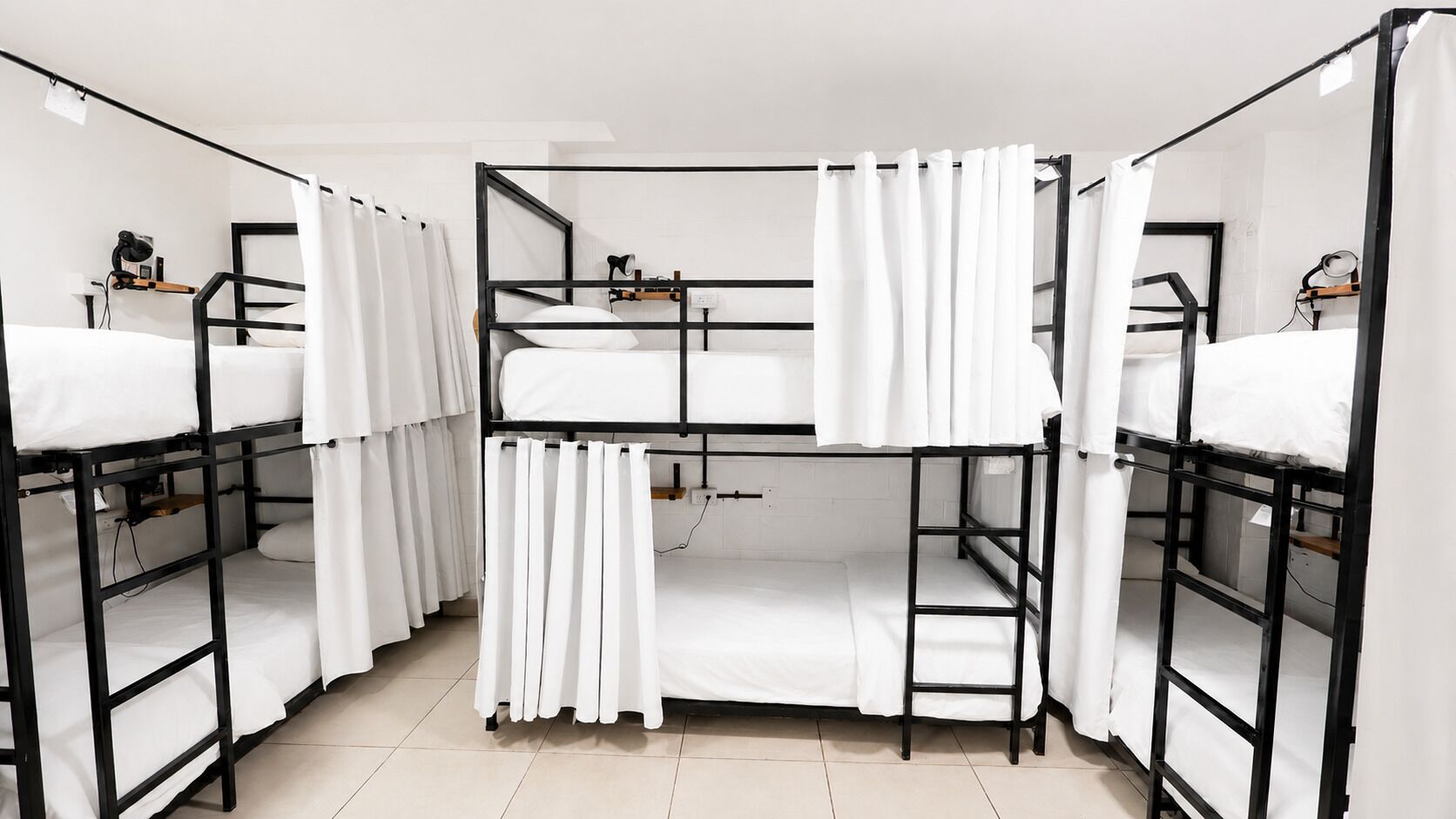 A dormitory room with bunk beds, white curtains, and a small shelf with a lamp.