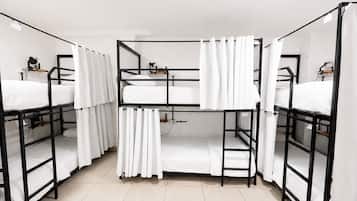 Um dormitório com beliches, cortinas brancas e um pequeno estante com uma luminária.