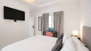 Quarto de hotel com cama, escrivaninha com laptop, televisão na parede e janela com cortinas.