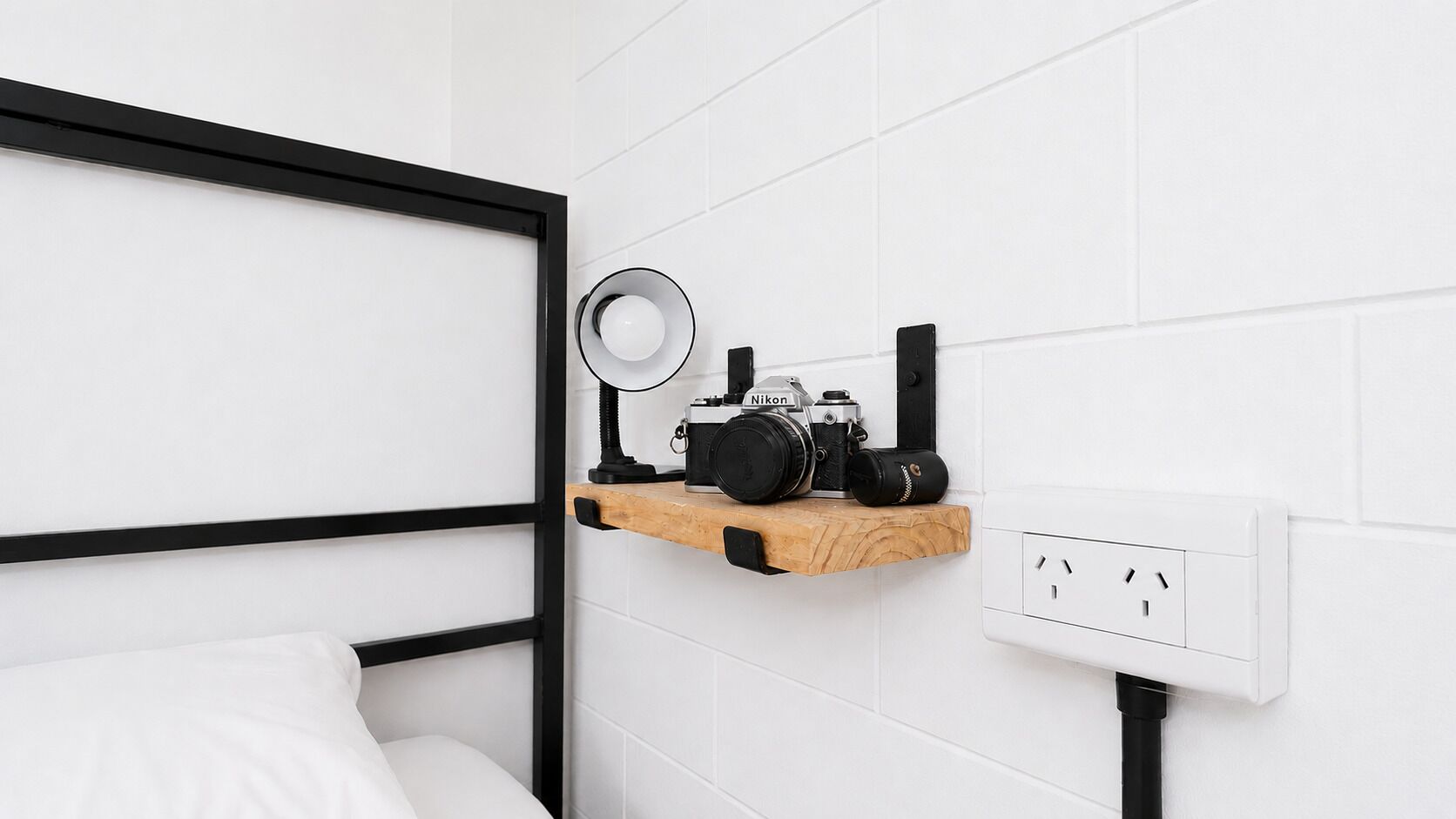 Un letto con struttura nera, uno scaffale di legno con una fotocamera e una lampada, e una parete bianca con una presa elettrica.