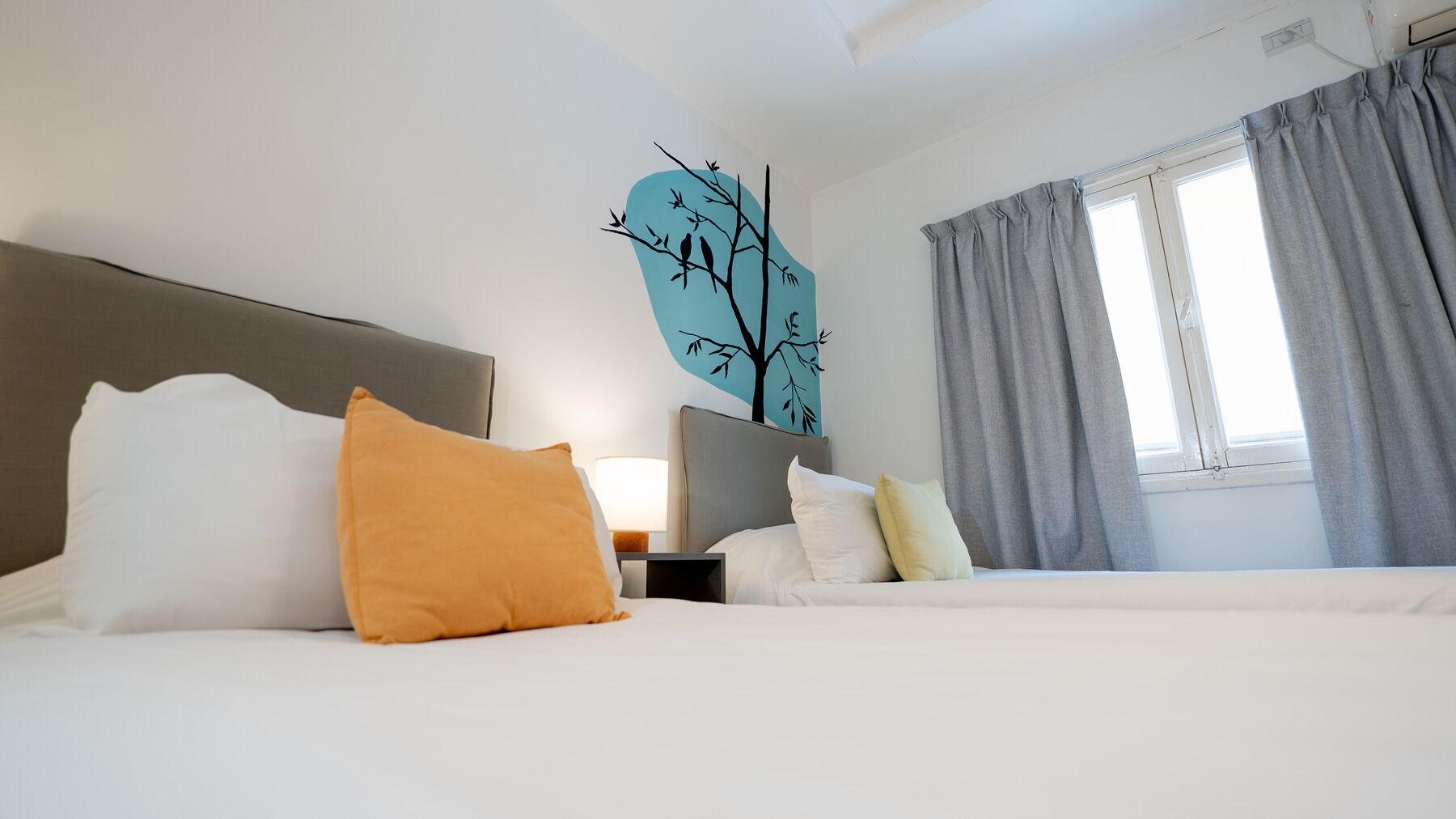 A hotel room with a bed, pillows, a nightstand, a lamp, a window with curtains, and a wall decoration of a tree.