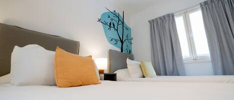A hotel room with a bed, pillows, a nightstand, a lamp, a window with curtains, and a wall decoration of a tree.