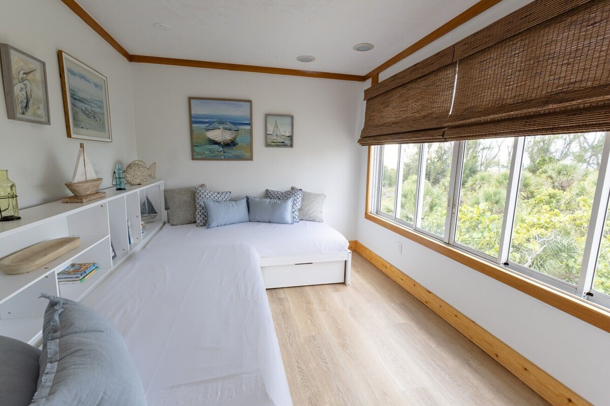 A bedroom with a large window, a bed, a shelf with decorative items, and wall art.
