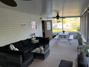 A covered patio area with a black sofa, wicker furniture, a glass coffee table, and a white table with chairs. - The Banyon House (Homosassa)