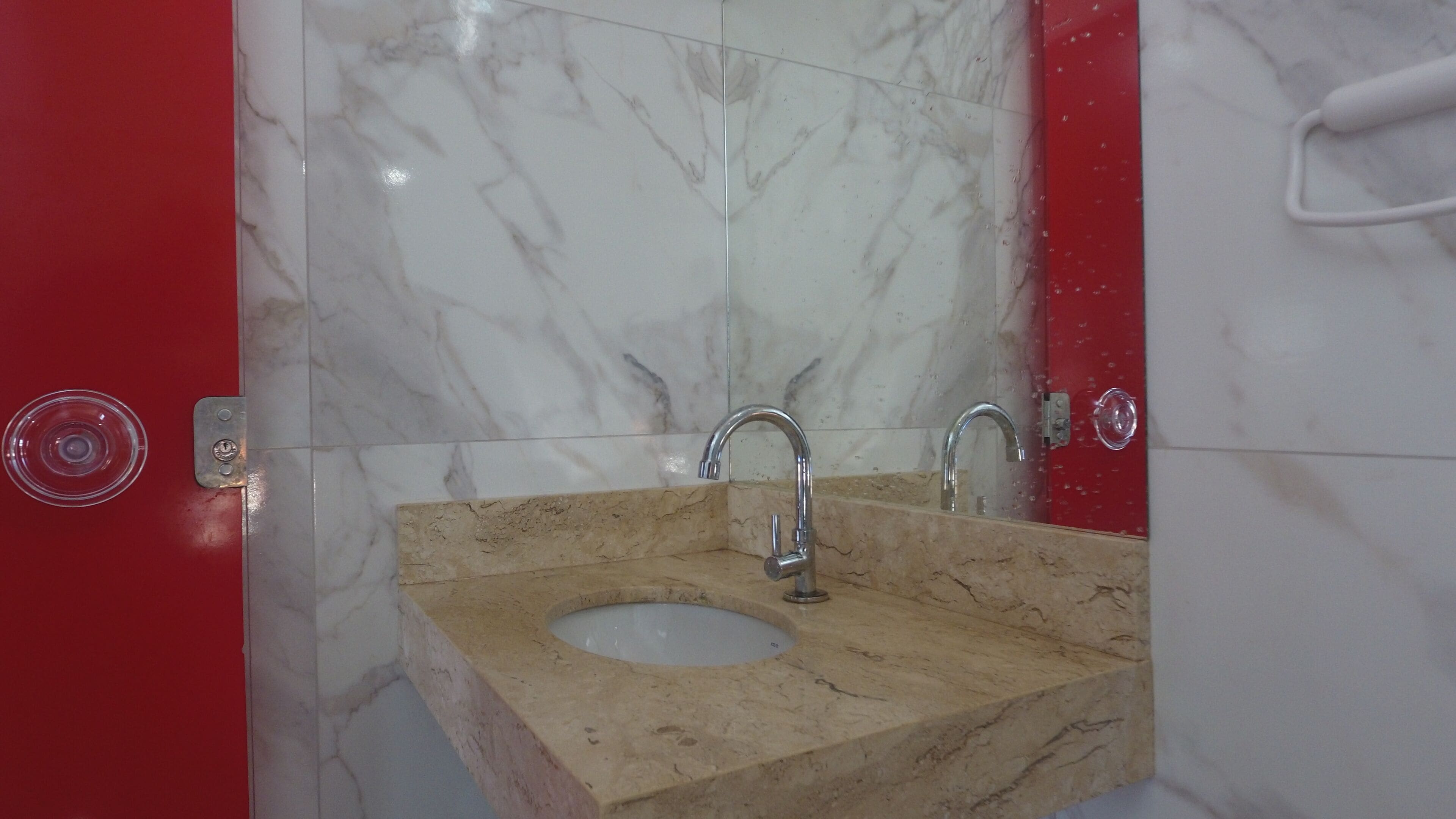 A bathroom with a marble sink and two faucets, a red door, and a mirror.