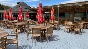 Eine Terrasse im Freien mit Holztischen und -stühlen, roten Sonnenschirmen und einer Holzdachkonstruktion.