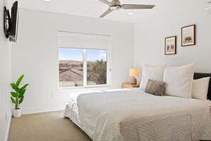 Un dormitorio con una cama grande, un ventilador de techo, una ventana con vistas a edificios, una mesita de noche con una lámpara y un cuadro en la pared.