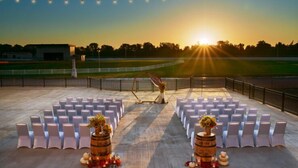 Un lieu de réception en plein air pour un mariage, avec des rangées de chaises blanches, des barils en bois décorés de fleurs et un coucher de soleil en arrière-plan.