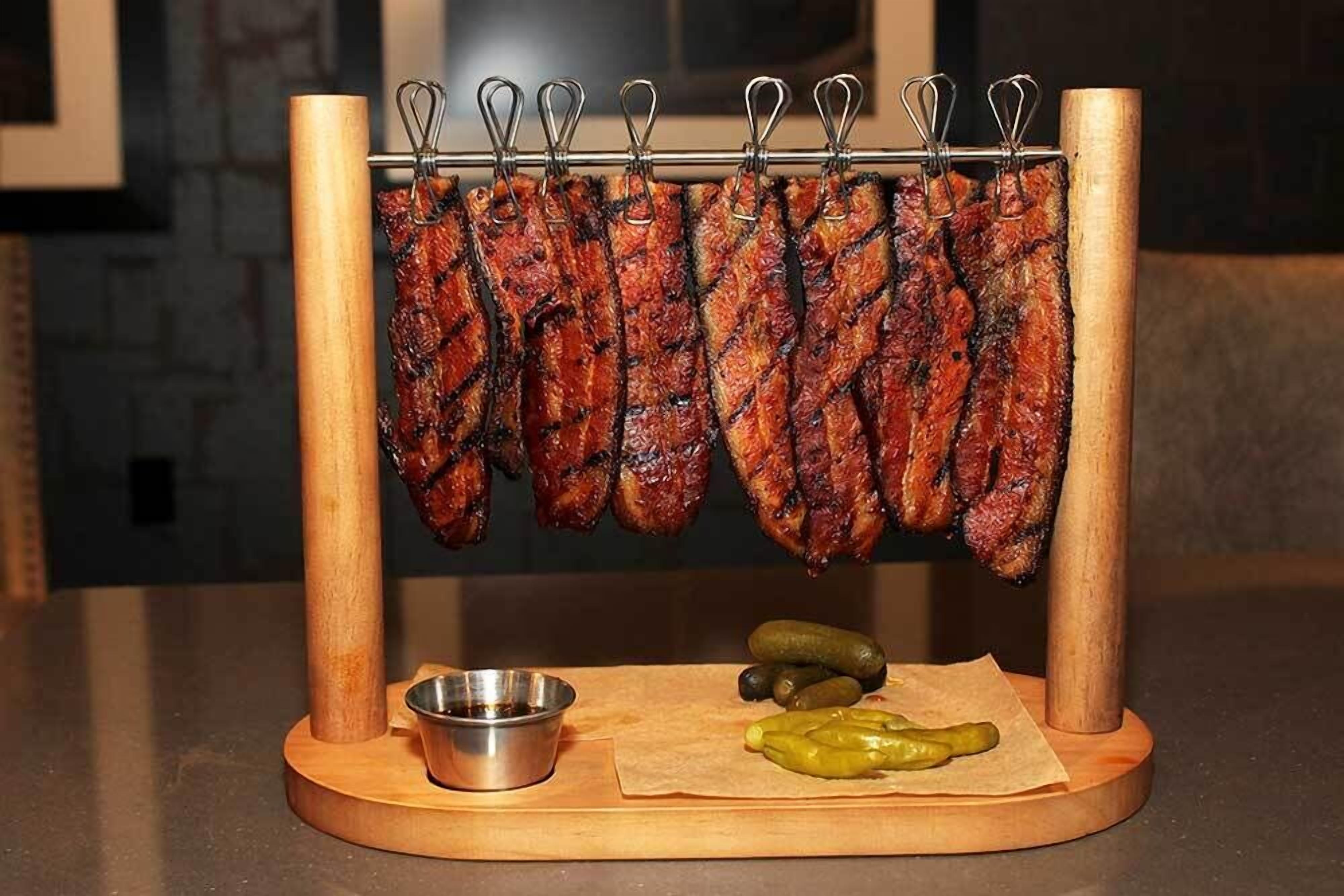 A wooden meat rack with grilled sausages and a small metal bowl and pickles on a wooden board.