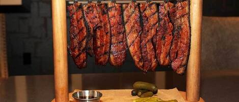 A wooden meat rack with grilled sausages and a small metal bowl and pickles on a wooden board.