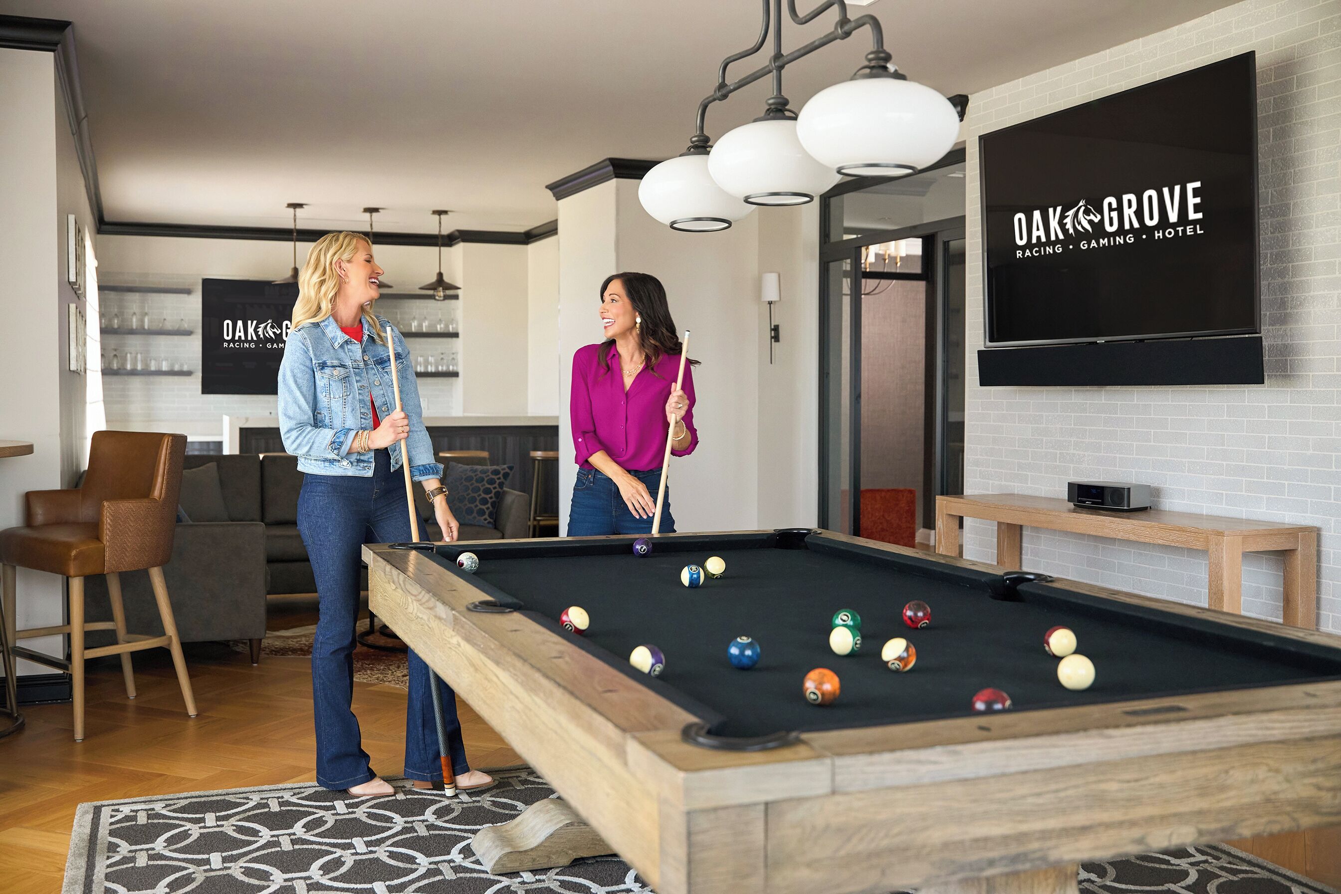 A pool table in a modern room with a brick wall and a mounted screen displaying 'OAK GROVE RACING GAMING HOTEL'.