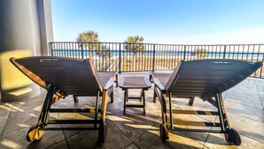A balcony with two lounge chairs and a small table, overlooking a beach with palm trees and a clear sky. - Where the Beach meets luxury! BEACHFRONT! (Pensacola)