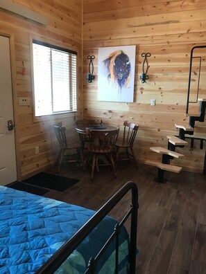A wooden cabin interior with a bed, a dining table with chairs, a painting on the wall, and a staircase.