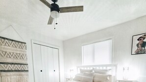 Un dormitori amb llit, ventilador de sostre, finestra amb persianes, estova amb dibuixos i un mur amb un element decoratiu.