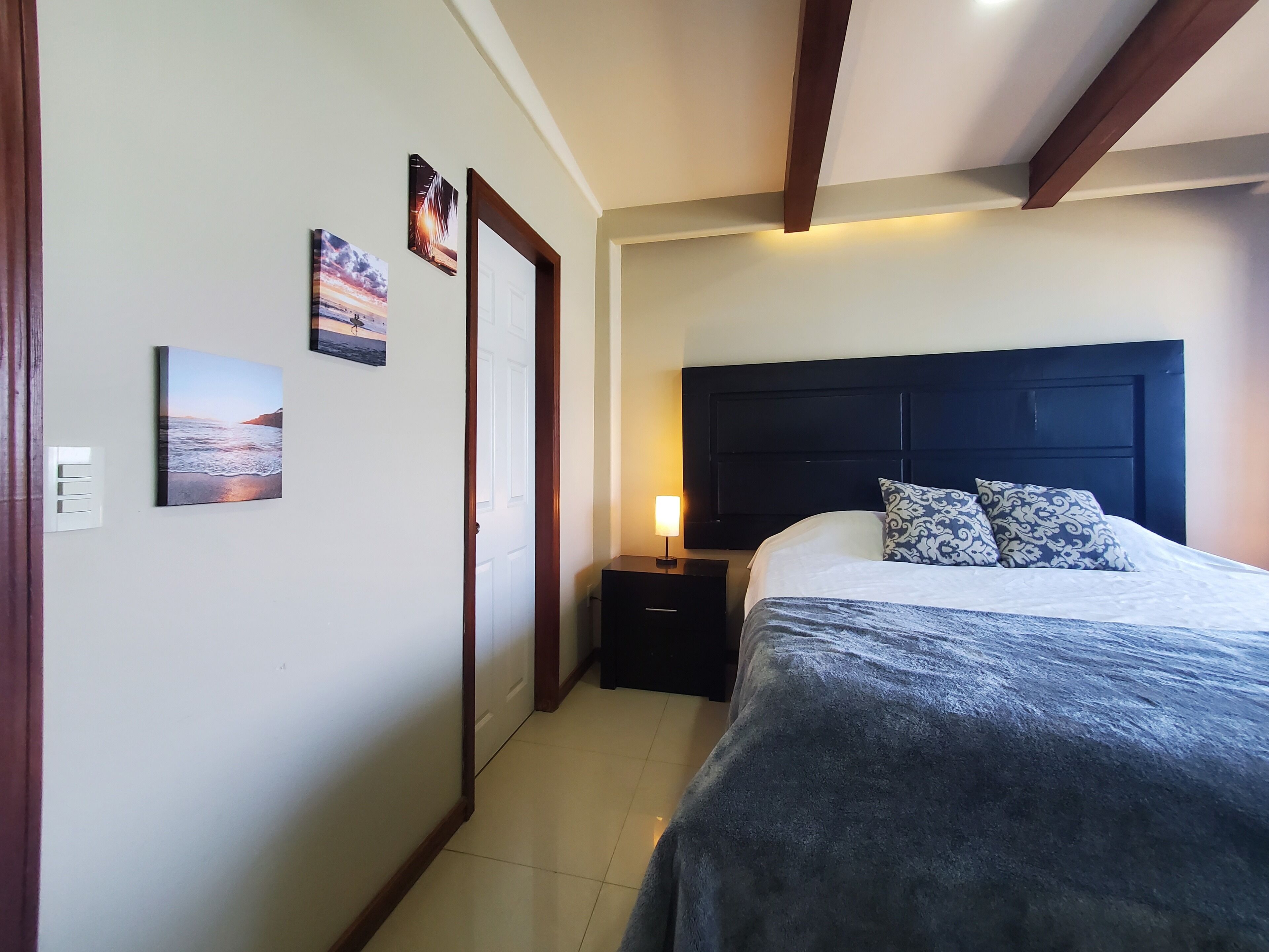 A bedroom with a bed, nightstand, and wall-mounted lamp. There are three framed pictures on the wall.