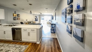 A modern kitchen with a central island, white cabinetry, and a dishwasher. There are multiple ceiling lights and a ceiling fan. The kitchen leads to a dining area with a table and chairs.