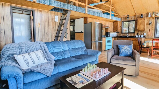 Un acogedor interior de cabaña con un sofá azul, una mesa de comedor de madera y una zona de cocina con refrigerador y estufa.