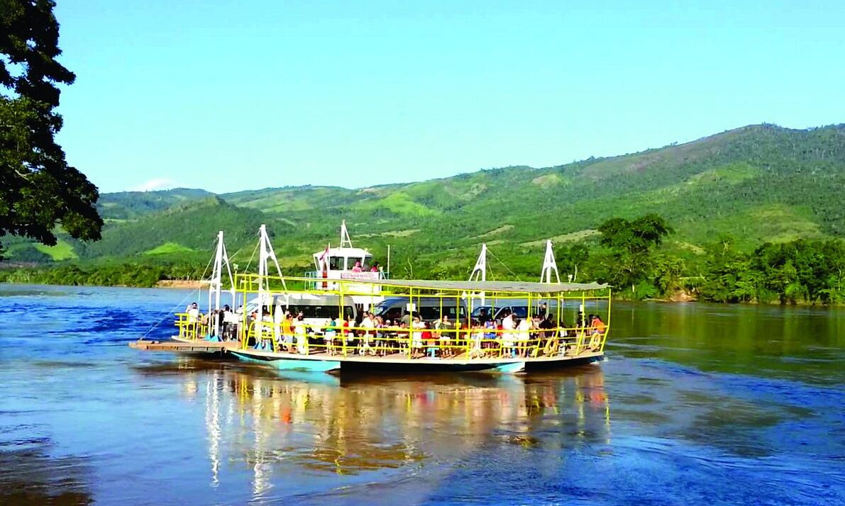 A riverboat with multiple decks and passengers, surrounded by lush green hills.