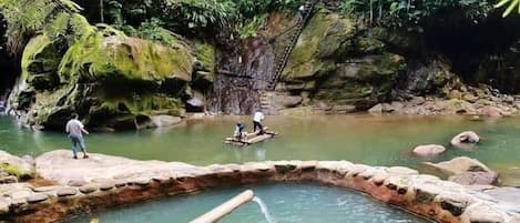 Una piscina natural rodeada de rocas y vegetación, con una pasarela de madera y una persona al borde.