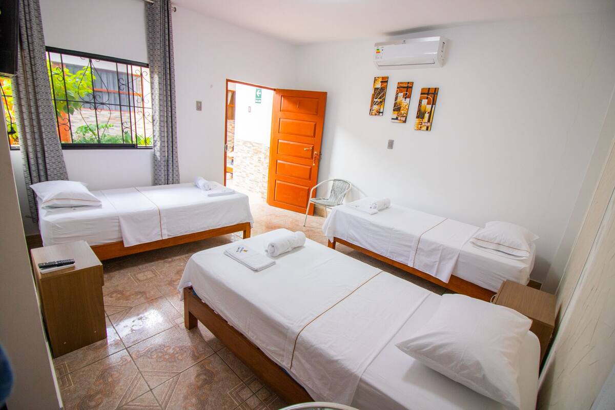 A hotel room with three single beds, a wooden nightstand, a small table, and a chair. There is a window with curtains, a door, and a wall-mounted air conditioner.