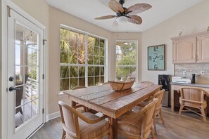 Un comedor con mesa de madera y sillas de mimbre, ventilador de techo y ventanas amplias con vistas a follaje.